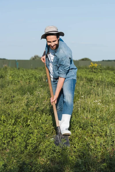 Vue pleine longueur de fermier heureux en chapeau bord et vêtements en denim creuser dans le champ — Photo de stock