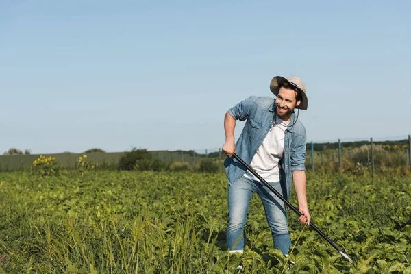 Улыбающийся фермер в шляпе смотрит в сторону, работая на зеленом поле — стоковое фото