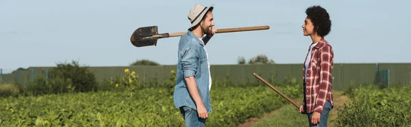 Agricultores multiétnicos alegres com ferramentas de jardinagem olhando uns para os outros no campo, bandeira — Fotografia de Stock