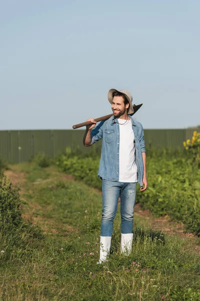 Ganzkörperansicht eines jungen Bauern mit Schaufel, der im Feld lächelt — Stockfoto