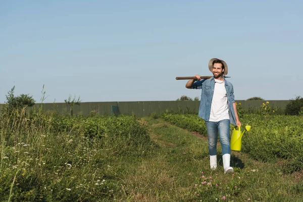 Ganzkörperansicht positiver Bauer mit Schaufel und Gießkanne, der im Feld spaziert — Stockfoto