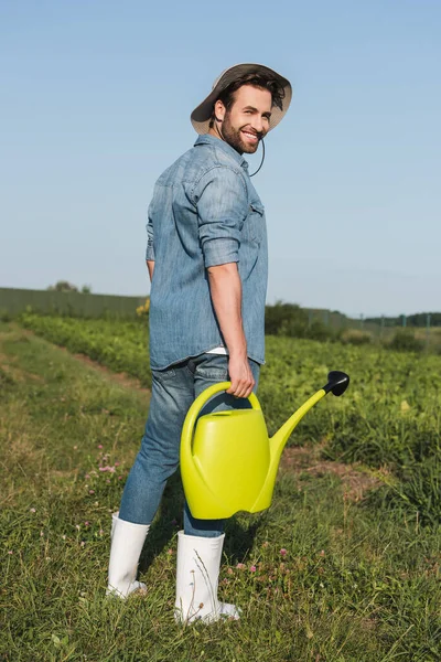 Vista de longitud completa de agricultor joven feliz de pie con regadera en el campo - foto de stock