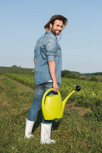 Ganzkörperansicht des glücklichen Jungbauern, der mit der Gießkanne auf dem Feld steht — Stockfoto