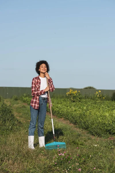 Vista completa de agricultor afroamericano feliz de pie con rastrillos en el campo - foto de stock