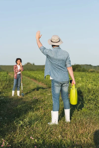 Farmer with watering can waving hand to blurred african american woman in field — Stock Photo