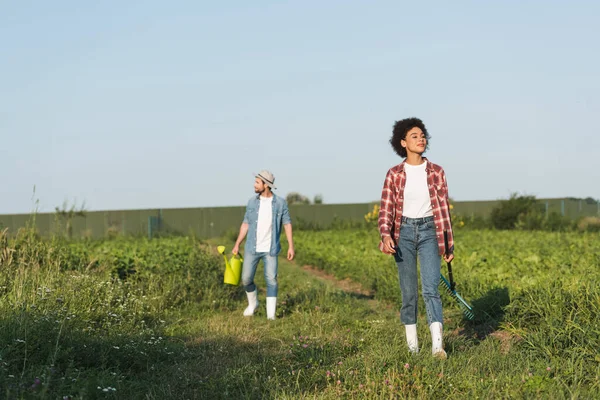 Agricultores multiétnicos con rastrillos y regaderas en tierras de cultivo bajo el cielo azul - foto de stock