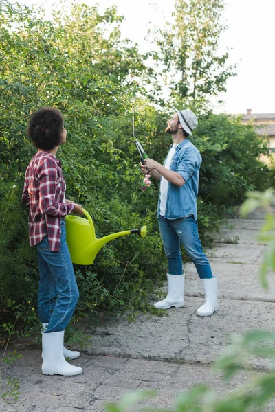 Afro-américaine femme tenant arrosoir peut près jardinier coupe buissons dans le jardin — Photo de stock