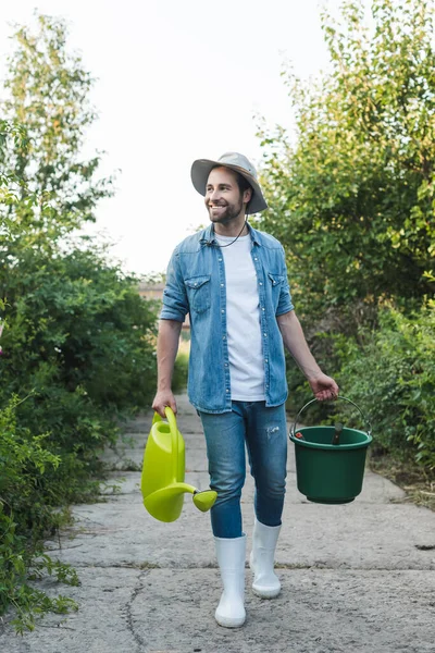 Vista completa de agricultor feliz con regadera y cubo caminando en el jardín - foto de stock