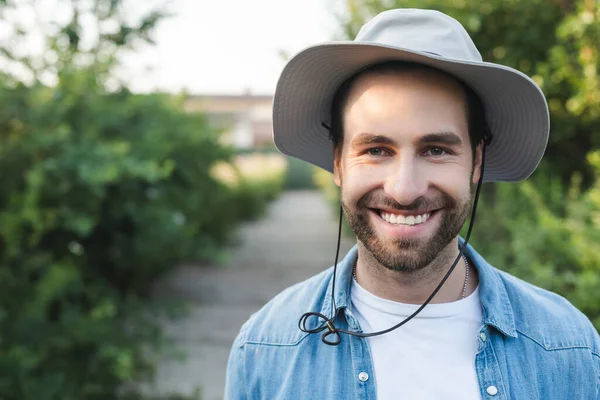 Joven y barbudo granjero en ala sombrero sonriendo a la cámara al aire libre - foto de stock