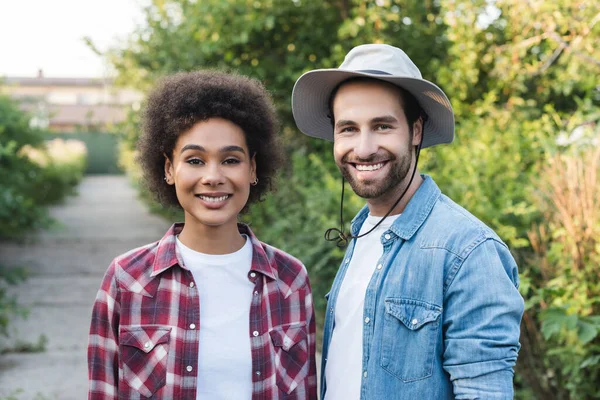 Jovens e felizes jardineiros inter-raciais olhando para a câmera no jardim borrado — Fotografia de Stock