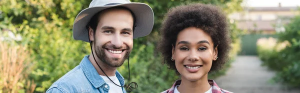 Agricultores multiétnicos alegres sorrindo para a câmera no jardim borrado, bandeira — Fotografia de Stock