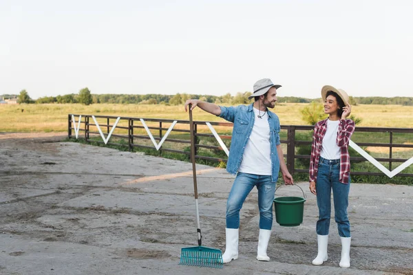 Happy farmer with rakes and bucket talking to young african american colleague — Stock Photo