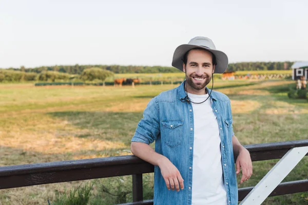 Contadino in cappello tesa e camicia di jeans in piedi vicino alla recinzione di legno e sorridente alla fotocamera — Foto stock