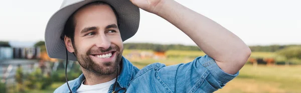 Положительный фермер в полях шляпы улыбаясь, глядя в сторону на открытом воздухе, баннер — стоковое фото