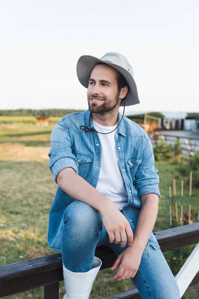 Giovane contadino sorridente in cappello tesa seduto su una recinzione di legno su terreni agricoli — Foto stock