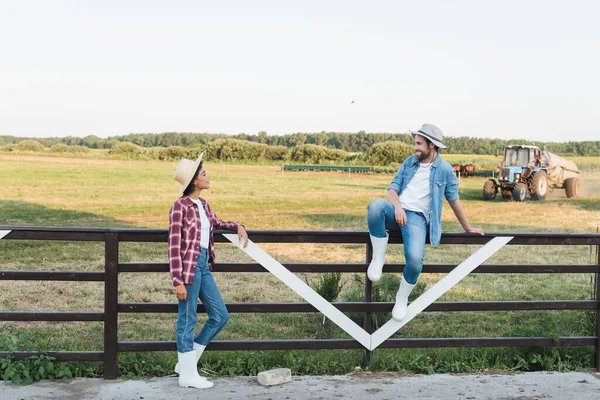 Ganzkörperansicht eines lächelnden Bauern auf einem Holzzaun in der Nähe eines jungen afrikanisch-amerikanischen Kollegen — Stockfoto