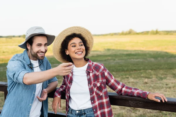 Farmer looking away and pointing with finger near happy african american colleague — Stock Photo