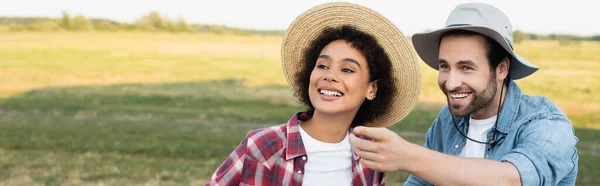 Agriculteur souriant regardant loin et pointant du doigt près de jeune femme afro-américaine, bannière — Photo de stock