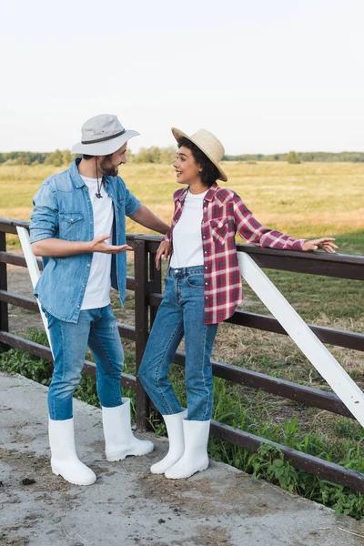 Vista completa de los agricultores interracial en sombreros hablando cerca de valla en la granja - foto de stock