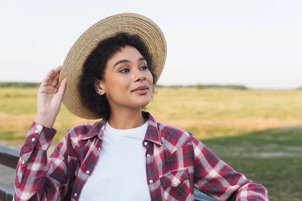 Pretty african american woman adjusting straw hat and looking away outdoors — Stock Photo