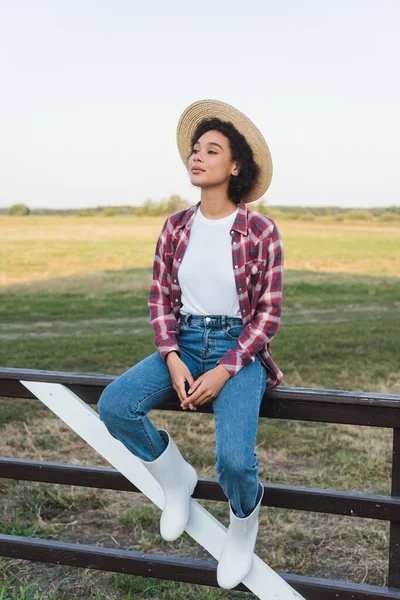 Full length view of african american woman in plaid shirt and jeans sitting on wooden fence — Stock Photo