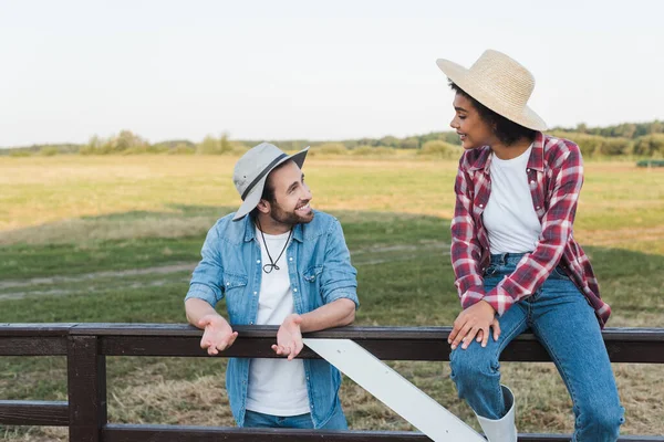 Granjero sonriente hablando con colega afroamericano sentado en valla en tierras de cultivo - foto de stock