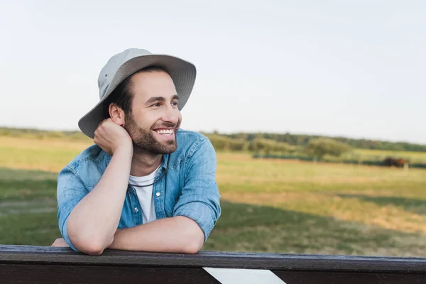 Agriculteur joyeux chapeau bord regardant loin près de la clôture dans les terres agricoles — Photo de stock