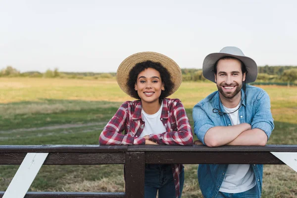 Alegre interracial agricultores em chapéus olhando para câmera perto de cerca de madeira no campo — Fotografia de Stock