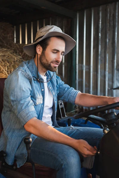 Agricultor barbudo con sombrero de ala y camisa de mezclilla sentado en el tractor en la granja - foto de stock