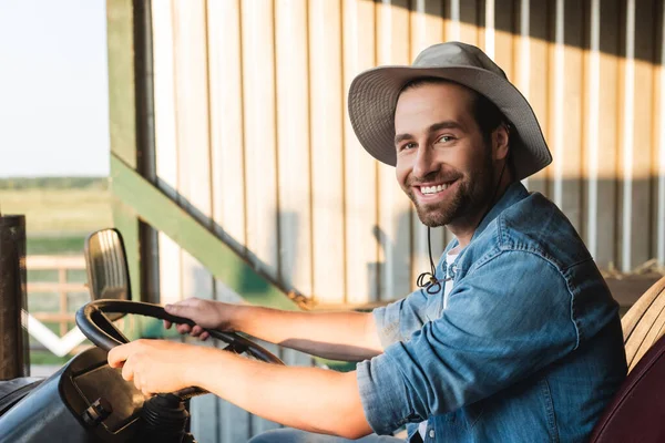 Heureux homme en chapeau bord regardant la caméra tout en étant assis sur le tracteur — Photo de stock