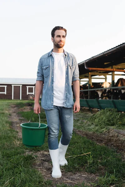 Full length view of farmer in denim clothes and rubber boots walking with bucket on farm — Stock Photo