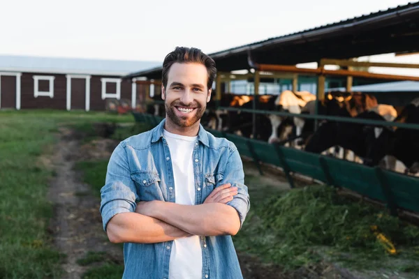 Agricultor feliz de pé com braços cruzados perto de vaqueiro borrado na fazenda — Fotografia de Stock