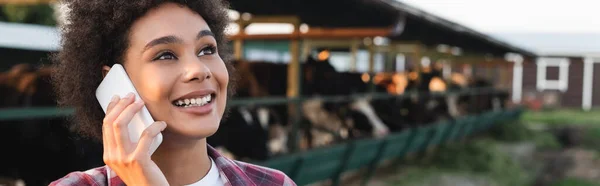 Heureuse femme afro-américaine parlant sur un téléphone mobile près de la maison de vache floue, bannière — Photo de stock