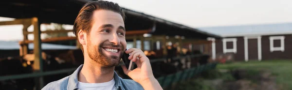 Granjero barbudo joven llamando en el teléfono inteligente cerca de granero borrosa, pancarta - foto de stock