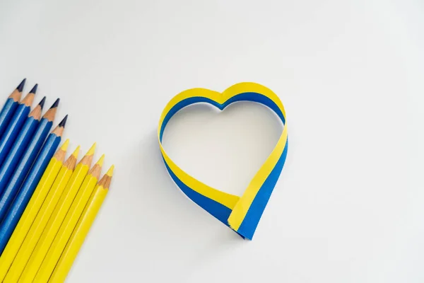 Vue du dessus du ruban bleu et jaune dans le signe de coeur près des crayons de couleur sur fond blanc — Photo de stock