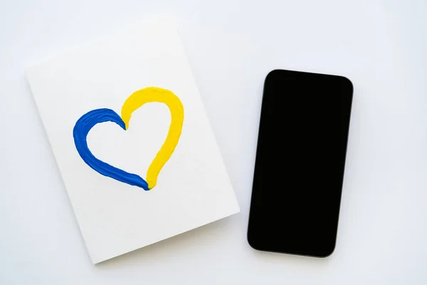 Vista dall'alto dello smartphone con schermo bianco vicino alla scheda con il segno del cuore blu e giallo dipinto su sfondo bianco — Foto stock