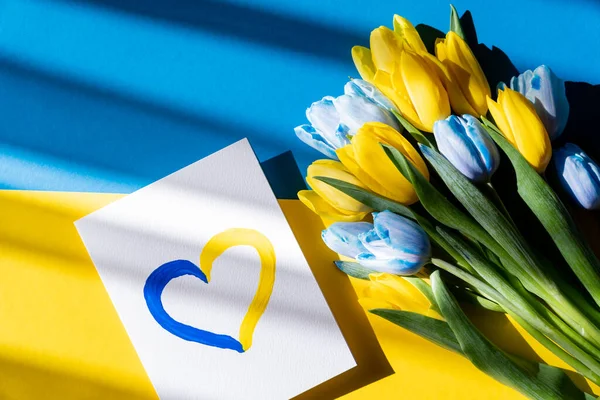 Top view of card with painted heart sign near blue and yellow tulips on ukrainian flag — Stock Photo