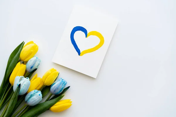 Top view of card with painted blue and yellow heart symbol near flowers on white background — Stock Photo