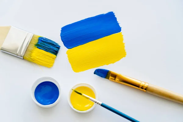 Vista superior de la bandera ucraniana pintada cerca de pinceles y pinturas sobre fondo blanco - foto de stock