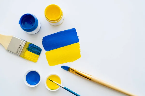 Draufsicht auf Farben und Pinsel in der Nähe der gemalten ukrainischen Flagge auf weißem Hintergrund — Stockfoto