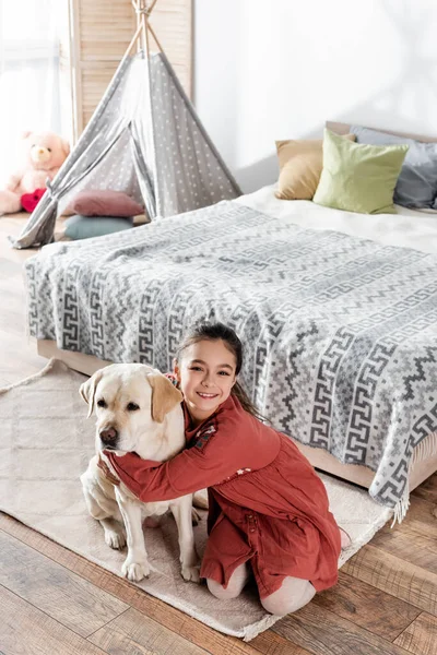 Visão de alto ângulo da menina feliz abraçando labrador perto da cama e wigwam — Fotografia de Stock