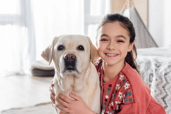 Aufgeregtes Mädchen blickt in die Kamera und umarmt gelben Labrador — Stockfoto