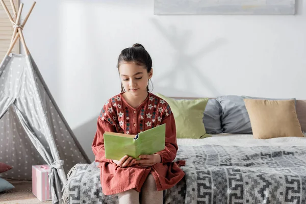 Preteen girl reading book while sitting on bed near pillows and wigwam on background — Stockfoto