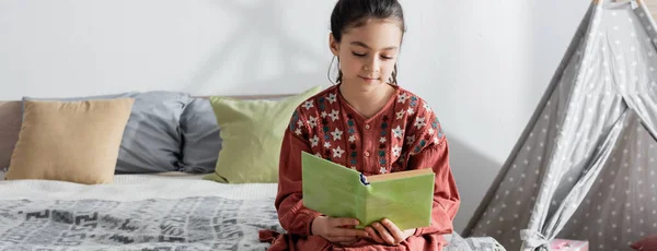 Preteen girl reading book while sitting on bed near pillows and wigwam, banner — Stockfoto