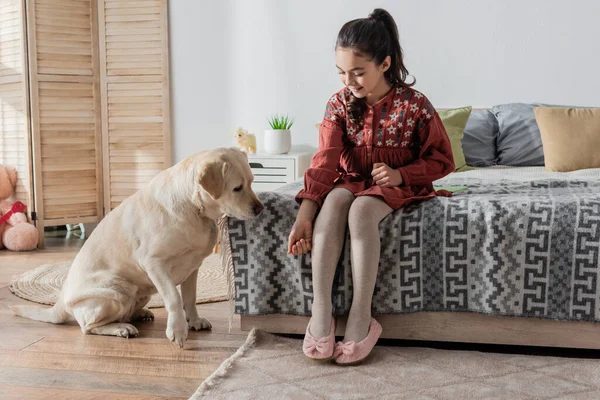 Full length view of smiling girl sitting on bed and playing with labrador — Stock Photo