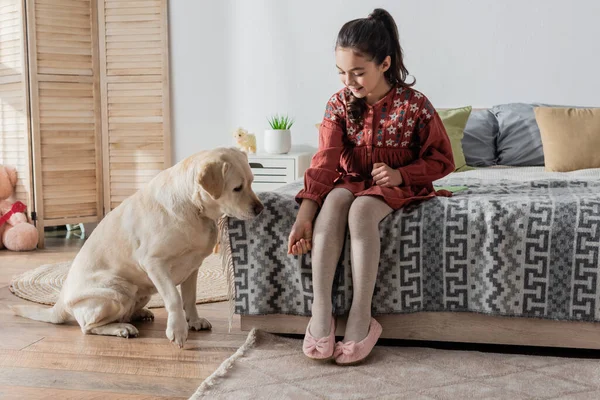 Piena vista lunghezza di sorridente ragazza seduta sul letto e giocare con labrador — Foto stock