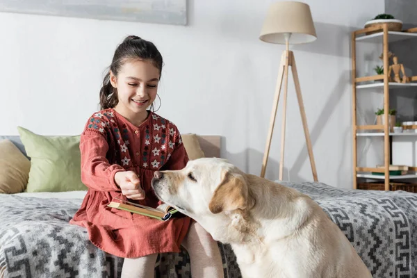 Sorrindo menina sentada no quarto com livro e brincando com labrador — Fotografia de Stock