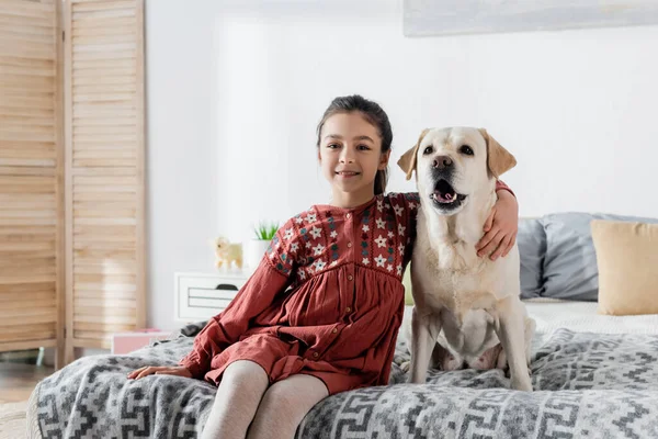 Glückliches brünettes Mädchen umarmt Labrador und lächelt in die Kamera, während es auf dem Bett sitzt — Stockfoto