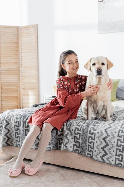 Happy preteen girl cuddling labrador dog while sitting on bed at home — Stock Photo