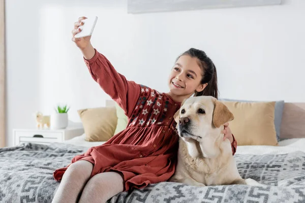 Menina satisfeito sentado na cama e tomando selfie com labrador — Fotografia de Stock